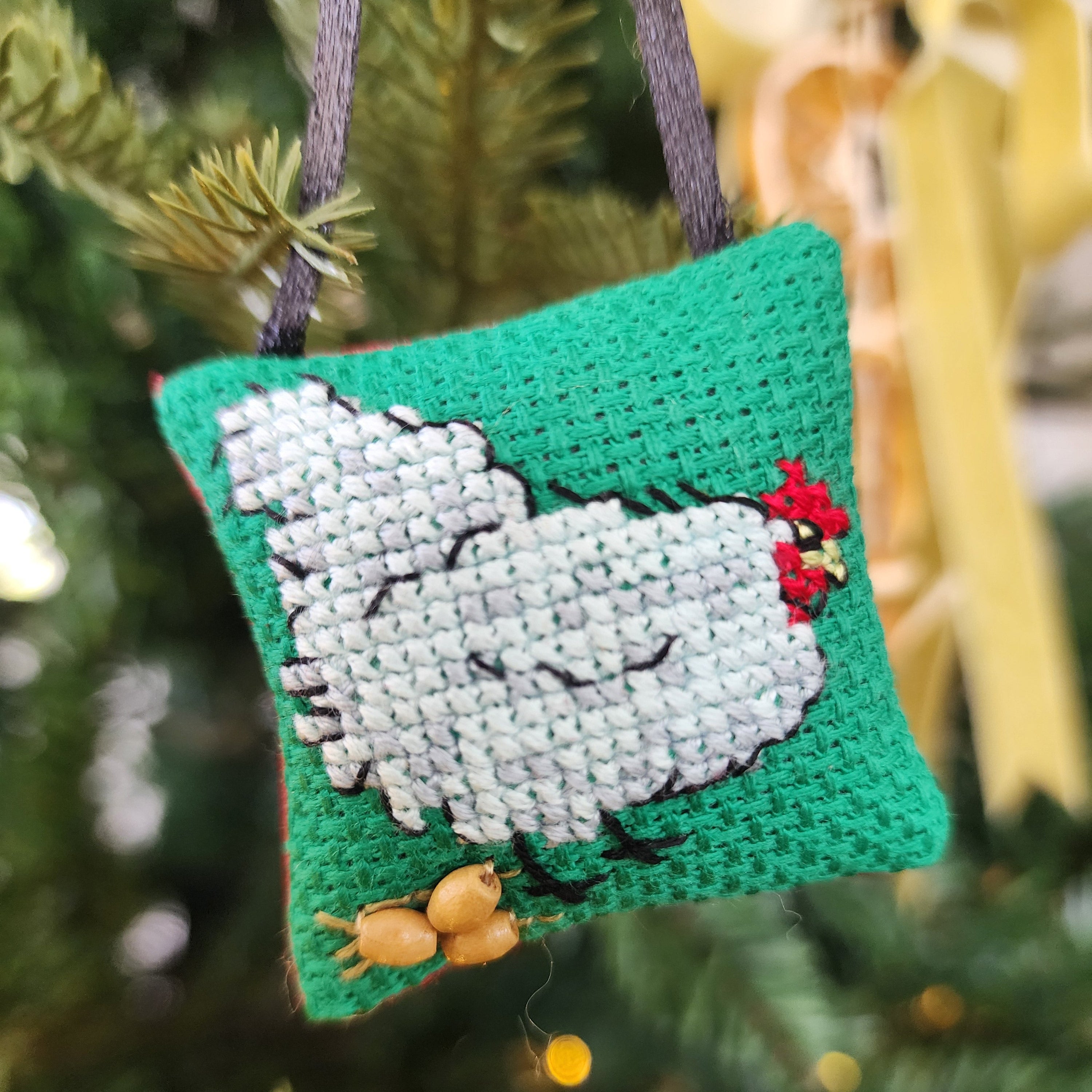 Cross-stitched ornament of a chicken laying eggs on green background, hanging on a Christmas tree.